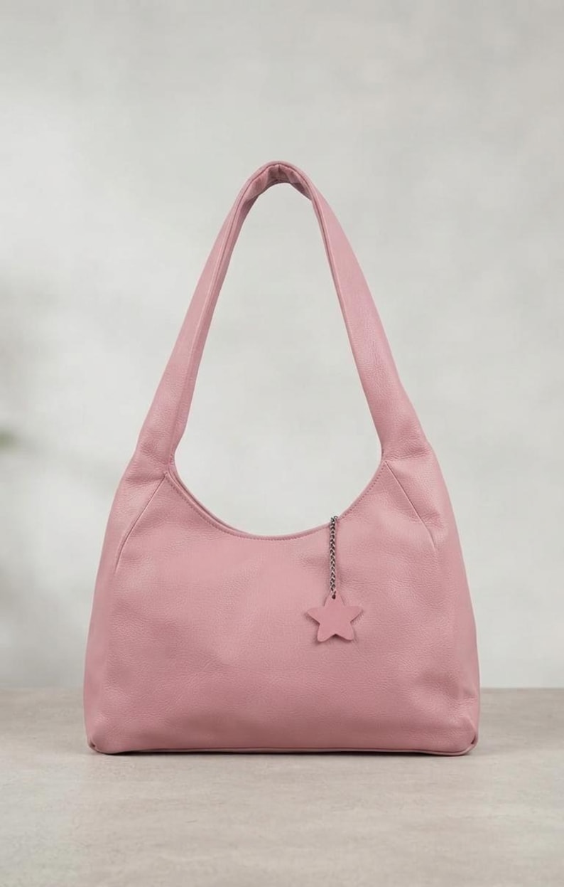 K&ouml;nnte beinhalten: Eine altrosa Hobo-Tasche mit glatter, matter Textur und einem einzelnen Schulterriemen. Die Tasche ist mit einem kleinen, f&uuml;nfzackigen Sternanh&auml;nger versehen, der an einer kurzen silberfarbenen Metallkette am oberen Rand befestigt ist.