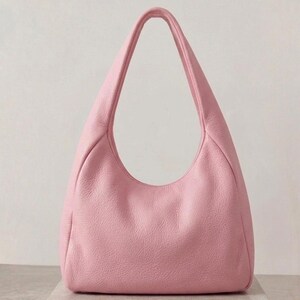 K&ouml;nnte beinhalten: Eine hellrosa Hobo-Tasche mit einer texturierten, genarbten Oberfl&auml;che. Die Tasche verf&uuml;gt &uuml;ber einen einzelnen integrierten Schulterriemen, der sich vom Hauptk&ouml;rper nach oben w&ouml;lbt und eine halbmondf&ouml;rmige Silhouette mit einer weiten &Ouml;ffnung bildet.