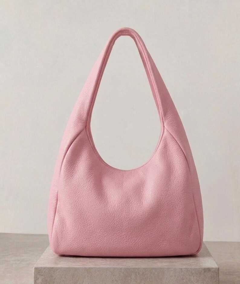 K&ouml;nnte beinhalten: Eine hellrosa Hobo-Tasche mit einer texturierten, genarbten Oberfl&auml;che. Die Tasche verf&uuml;gt &uuml;ber einen einzelnen integrierten Schulterriemen, der sich vom Hauptk&ouml;rper nach oben w&ouml;lbt und eine halbmondf&ouml;rmige Silhouette mit einer weiten &Ouml;ffnung bildet.