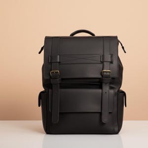 Peut inclure: Un sac à dos en cuir noir avec une poignée supérieure, des bretelles réglables et deux boucles avant. Le sac à dos a une fermeture à rabat et deux poches latérales. Le sac est posé sur une surface blanche.