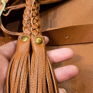 以下が含まれることがあります： タンカラーのシボ革製バッグアクセサリー。2本の編み込みストラップが長い細いフリンジタッセルにつながっています。各タッセルは上部で小さな丸い真鍮色の金属製リベットで固定されています。