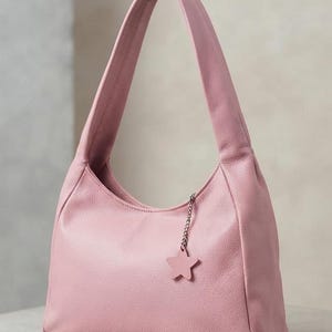 K&ouml;nnte beinhalten: Eine hellrosa Schultertasche im Hobo-Stil mit strukturierter Oberfl&auml;che. Die Tasche verf&uuml;gt &uuml;ber einen einzelnen breiten Trageriemen und eine kleine silberfarbene Metallkette am oberen Rand, an der ein passender sternf&ouml;rmiger Anh&auml;nger h&auml;ngt.