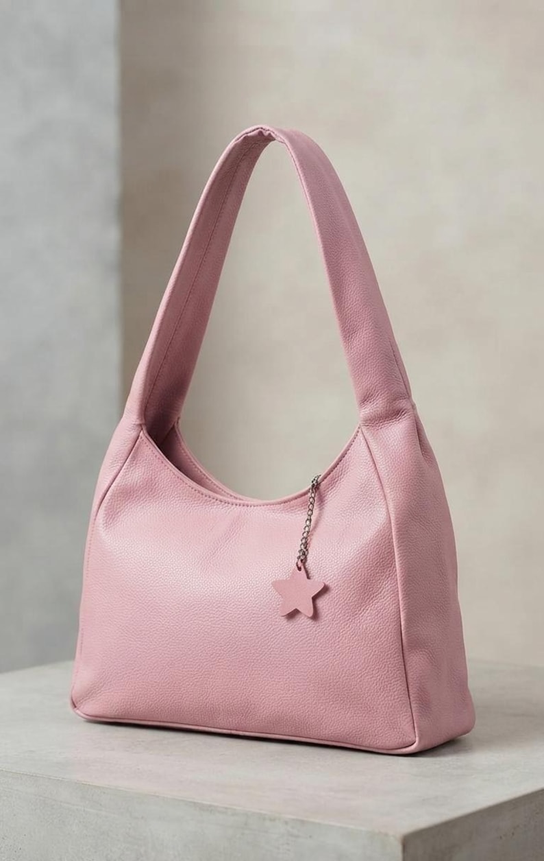 K&ouml;nnte beinhalten: Eine hellrosa Schultertasche im Hobo-Stil mit strukturierter Oberfl&auml;che. Die Tasche verf&uuml;gt &uuml;ber einen einzelnen breiten Trageriemen und eine kleine silberfarbene Metallkette am oberen Rand, an der ein passender sternf&ouml;rmiger Anh&auml;nger h&auml;ngt.