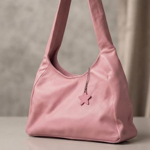 K&ouml;nnte beinhalten: Eine altrosa Umh&auml;ngetasche im Hobo-Stil mit einer weichen, genarbten Textur. Die Tasche verf&uuml;gt &uuml;ber einen einzelnen breiten Riemen und einen kleinen, passenden sternf&ouml;rmigen Anh&auml;nger in Rosa an einer d&uuml;nnen silberfarbenen Metallkette.