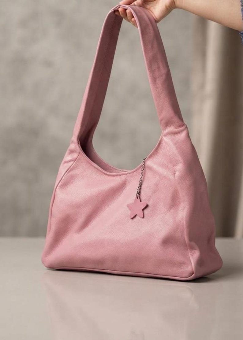K&ouml;nnte beinhalten: Eine altrosa Umh&auml;ngetasche im Hobo-Stil mit einer weichen, genarbten Textur. Die Tasche verf&uuml;gt &uuml;ber einen einzelnen breiten Riemen und einen kleinen, passenden sternf&ouml;rmigen Anh&auml;nger in Rosa an einer d&uuml;nnen silberfarbenen Metallkette.