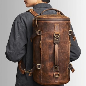 Peut inclure: Sac à dos en cuir marron de forme cylindrique. Il est doté d'une fermeture éclair sur le dessus, de bretelles réglables et d'éléments métalliques. Le cuir présente une finition vieillie, lui donnant un aspect vintage. Le sac est conçu pour être porté sur le dos.