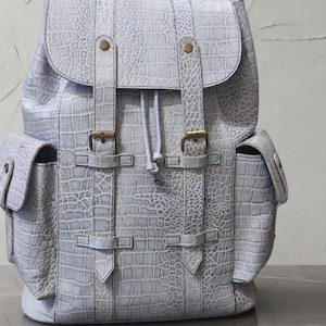 Puede incluir: Una mochila de color gris claro con un patrón texturizado de piel de cocodrilo. La bolsa incluye una solapa superior con dos correas verticales, hebillas metálicas de color latón y dos bolsillos laterales. Un cierre de cordón asegura el compartimento.
