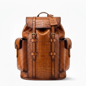 Puede incluir: Una mochila de cuero marrón con un patrón texturizado de piel de cocodrilo. La bolsa cuenta con dos bolsillos laterales, un asa superior y una solapa frontal asegurada por dos correas verticales con hebillas metálicas. Una etiqueta dice Handmade Est 2022.