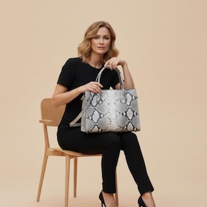 Könnte beinhalten: Grau-weiß gemusterte Tragetasche mit Schlangenmuster und strukturiertem Design. Die Tasche hat zwei obere Griffe und einen Schultergurt. Die Tasche wird von einer Person in einem schwarzen Outfit und schwarzen Absätzen gehalten. Der Hintergrund ist einfarbig beige.