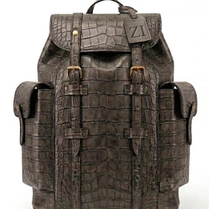 May include: A dark grey backpack with a textured crocodile skin pattern. It features a top flap with two vertical straps, brass-toned buckles, and two side pockets with snap closures. A rectangular luggage tag with the letters ZI is attached.
