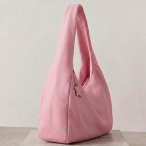 K&ouml;nnte beinhalten: Eine zartrosa Hobo-Tasche mit strukturierter Oberfl&auml;che und einem einzelnen Schulterriemen. Die Tasche verf&uuml;gt &uuml;ber einen kleinen sternf&ouml;rmigen Anh&auml;nger an einer silberfarbenen Kette nahe dem Rei&szlig;verschluss an der Oberkante.