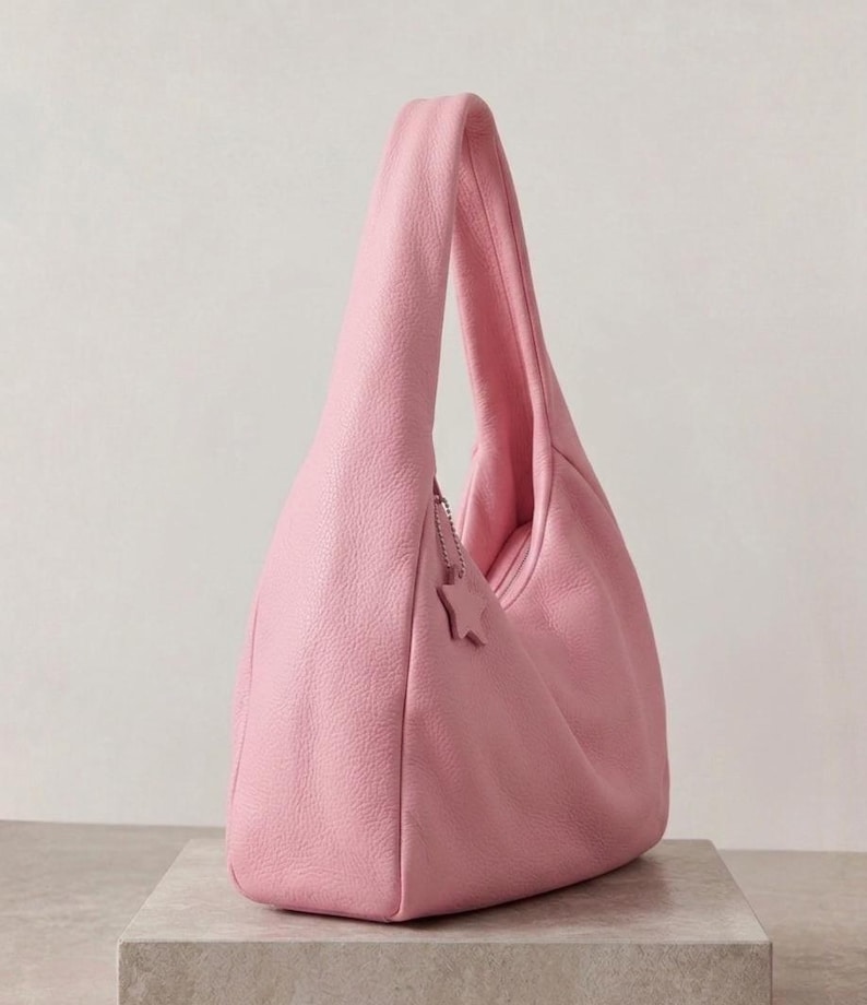 K&ouml;nnte beinhalten: Eine zartrosa Hobo-Tasche mit strukturierter Oberfl&auml;che und einem einzelnen Schulterriemen. Die Tasche verf&uuml;gt &uuml;ber einen kleinen sternf&ouml;rmigen Anh&auml;nger an einer silberfarbenen Kette nahe dem Rei&szlig;verschluss an der Oberkante.