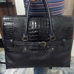 May include: A black briefcase featuring a glossy crocodile-embossed leather upper section and a textured pebbled leather lower section. The bag includes two top handles, a horizontal buckled strap across the centre, and gold-tone metal hardware.