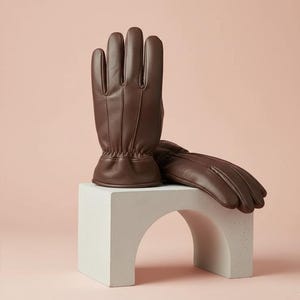 以下が含まれることがあります： 茶色の革手袋のペア。1つは直立し、もう1つは白いアーチ型の台座に置かれています。手袋は、ステッチが見え、表面にテクスチャ加工が施されたクラシックなデザインです。背景はソフトピンクです。