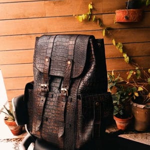 Könnte beinhalten: Ein dunkelbrauner Rucksack mit einer strukturierten Krokodilleder-Optik. Die Tasche verfügt über eine obere Klappe, die mit zwei vertikalen Riemen und Metallschnallen gesichert ist, sowie Seitentaschen mit Druckknöpfen. Strukturierte Form.