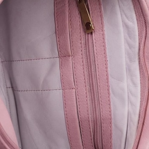 K&ouml;nnte beinhalten: Das Innere einer hellrosa Ledertasche mit einer vertikalen Rei&szlig;verschlusstasche und einem goldfarbenen Metallzipper. Das Innenfutter ist in einem helleren Pastellrosa gehalten, und die Tasche ist mit passendem rosa Leder eingefasst.