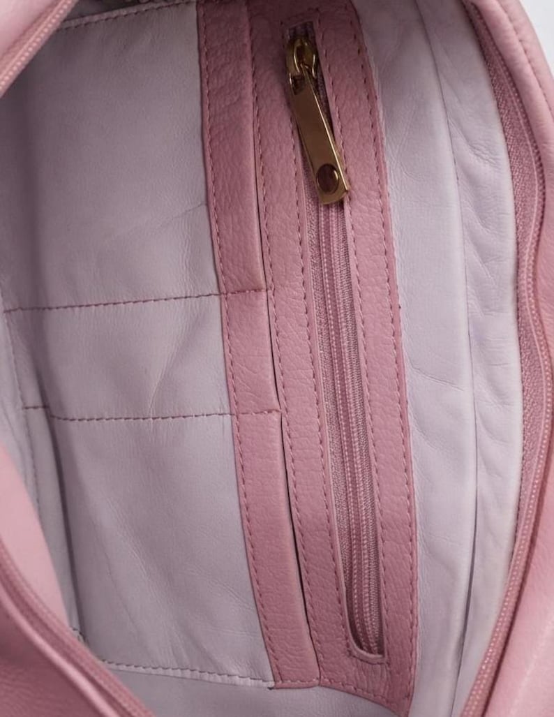 K&ouml;nnte beinhalten: Das Innere einer hellrosa Ledertasche mit einer vertikalen Rei&szlig;verschlusstasche und einem goldfarbenen Metallzipper. Das Innenfutter ist in einem helleren Pastellrosa gehalten, und die Tasche ist mit passendem rosa Leder eingefasst.