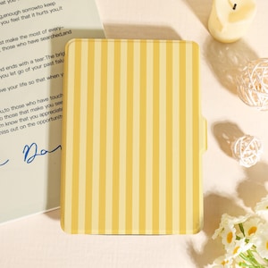 May include: A yellow and cream striped notebook cover with a tab on the right side. The notebook cover is rectangular and has vertical stripes. A handwritten note is partially visible in the background.