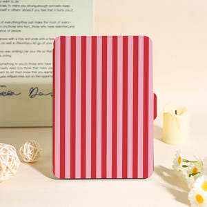 May include: A rectangular case featuring a vertical striped design in alternating red and pink. The case has a small tab on the side. The background includes a candle, decorative balls, and flowers.
