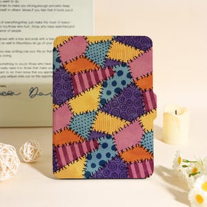 May include: A rectangular e-reader cover featuring a colorful patchwork design of yellow, purple, pink, and blue fabric segments. The pattern includes various textures like stripes, swirls, and dots, all joined by black stitched X-shaped details.