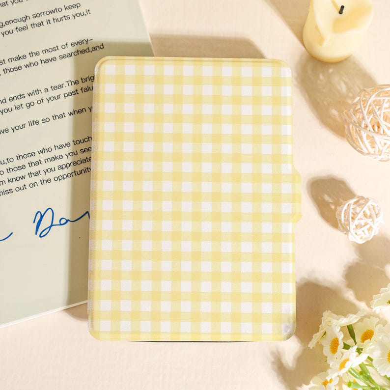 May include: A yellow and white gingham pattern case, possibly for a tablet or e-reader, with a rounded shape and a clasp. The case is set against a soft, neutral background with a handwritten note and decorative elements.