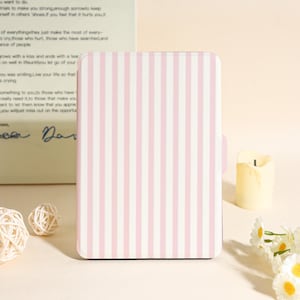 Op de afbeelding: Een rechthoekige, roze en wit gestreepte notebook- of tablethoes. De hoes heeft een verticaal streepjespatroon en een afgeronde bovenrand. Aan de rechterkant is een klein lipje zichtbaar. De achtergrond omvat een kaars, bloemen en een handgeschreven notitie.