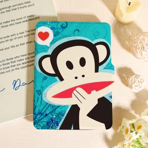 May include: A teal phone case featuring a cartoon monkey with a red mouth and a heart-shaped speech bubble. The case has a white background with blue swirls and the monkey is holding its hand up to its mouth.