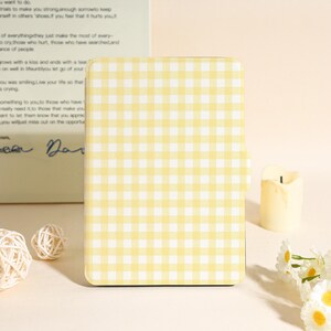 May include: A yellow and white gingham pattern phone case. The case is rectangular with rounded corners. The background includes a candle, decorative flowers, and a book with handwritten text.