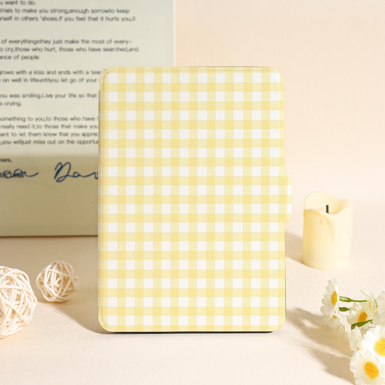 May include: A yellow and white gingham pattern phone case. The case is rectangular with rounded corners. The background includes a candle, decorative flowers, and a book with handwritten text.