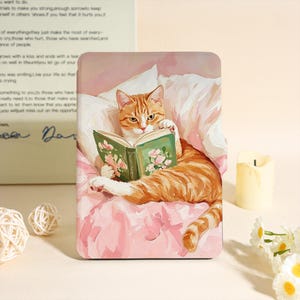 Puede incluir: Una funda rectangular con un gato atigrado jengibre leyendo un libro con un diseño floral. El gato está acostado sobre una superficie rosa con una almohada blanca. El libro tiene una cubierta verde y detalles florales rosas.
