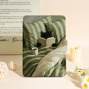 May include: A decorative print showing a black cat engrossed in a book while in bed, with green and white striped bedding. A white mug with coffee is placed nearby. A candle and flowers are also in the image.