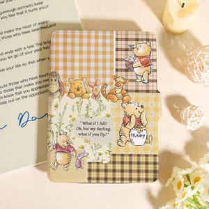 Può includere: Una custodia decorativa con Winnie the Pooh e i suoi amici. Il design incorpora vari motivi, tra cui il vichy giallo e il tartan. La custodia presenta illustrazioni di Pooh, Pimpi, Tigro e Eeyore. Il testo sulla custodia recita: "What if I fall? Oh, but my darling, what if you fly?"
