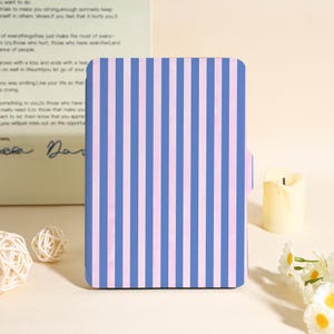 Può includere: Una custodia rettangolare con un motivo a righe blu e rosa. La custodia ha una piccola linguetta laterale. Lo sfondo include una candela, fiori e testo di un libro.