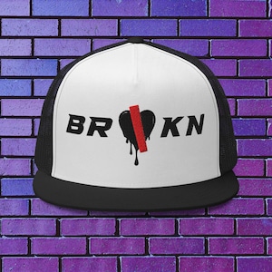 Puede incluir: Gorra de camionero en blanco y negro con la palabra "BRKN" en letras negras. Un corazón negro con una franja roja está entre las letras, con gotas negras. La gorra tiene una visera negra y una parte trasera de malla.