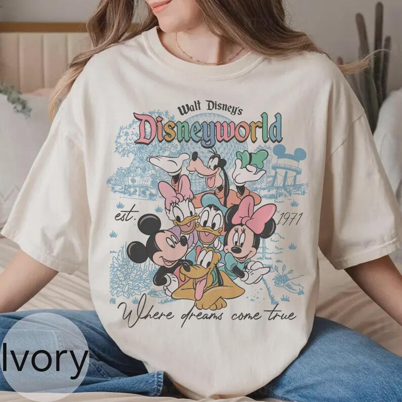 Op de afbeelding: Ivoor kleurig T-shirt met een kleurrijke Disney World afbeelding. Het ontwerp bevat Mickey Mouse, Minnie Mouse, Donald Duck, Goofy en Pluto. Tekst bevat "Walt Disney's Disneyworld", "est. 1971" en "Where dreams come true".