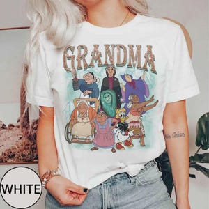 Puede incluir: Camiseta blanca con la palabra "GRANDMA" en una fuente de estilo retro, sobre un gr&aacute;fico colorido de abuelas de dibujos animados. La camiseta tiene mangas cortas y cuello redondo.