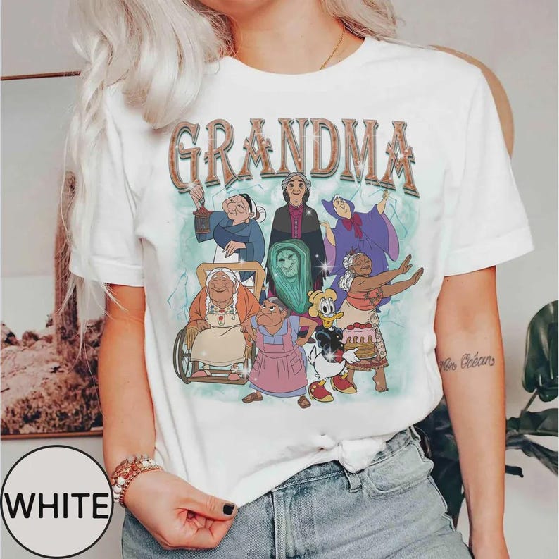 Puede incluir: Camiseta blanca con la palabra "GRANDMA" en una fuente de estilo retro, sobre un gr&aacute;fico colorido de abuelas de dibujos animados. La camiseta tiene mangas cortas y cuello redondo.
