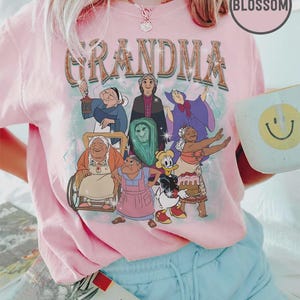 Puede incluir: Camiseta rosa claro con la palabra "GRANDMA" en una fuente vintage, con abuelas de dibujos animados. La camiseta tiene cuello redondo y mangas cortas. Tambi&eacute;n se ve una taza blanca con una carita sonriente amarilla y un libro.