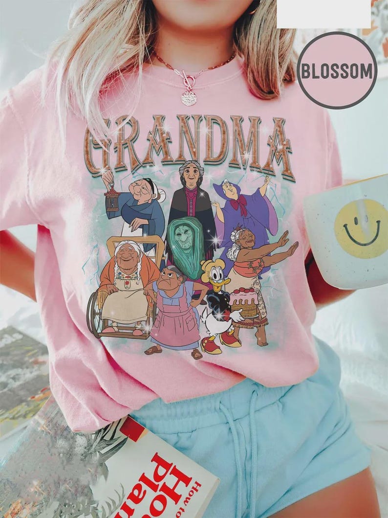 Puede incluir: Camiseta rosa claro con la palabra "GRANDMA" en una fuente vintage, con abuelas de dibujos animados. La camiseta tiene cuello redondo y mangas cortas. Tambi&eacute;n se ve una taza blanca con una carita sonriente amarilla y un libro.