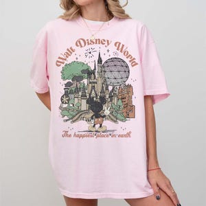Puede incluir: Una camiseta rosa claro con un gr&aacute;fico de estilo vintage de Walt Disney World. El dise&ntilde;o incluye un castillo, Spaceship Earth, Mickey Mouse y el texto "The happiest place on earth."