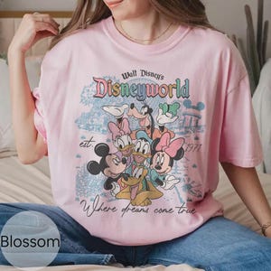 Op de afbeelding: Lichtroze T-shirt met een Disney World-afbeelding met Mickey Mouse, Minnie Mouse, Donald Duck, Katrien Duck, Goofy en Pluto. Het shirt heeft de tekst "Walt Disney's Disneyworld" en "Where dreams come true."