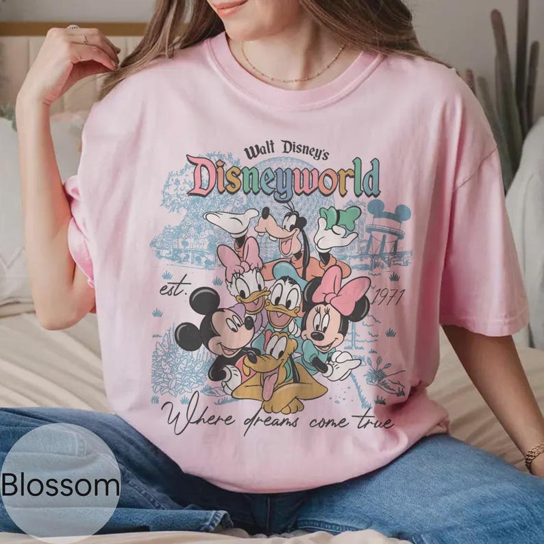 Op de afbeelding: Lichtroze T-shirt met een Disney World-afbeelding met Mickey Mouse, Minnie Mouse, Donald Duck, Katrien Duck, Goofy en Pluto. Het shirt heeft de tekst "Walt Disney's Disneyworld" en "Where dreams come true."