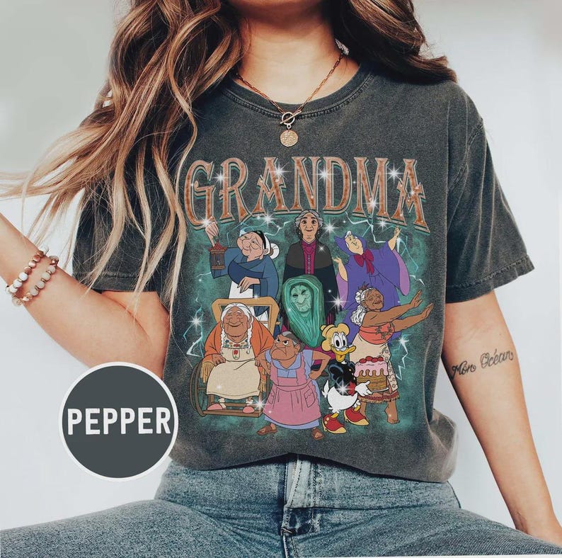 Puede incluir: Camiseta gris oscuro con la palabra "GRANDMA" en un estilo vintage. La camiseta presenta un gr&aacute;fico de abuelas de dibujos animados sobre un fondo verde y turquesa. La camiseta es de cuello redondo.