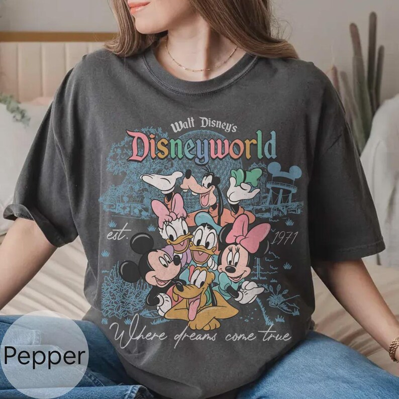 Op de afbeelding: Donkergrijs t-shirt met een kleurrijk Disney World-ontwerp met Mickey Mouse, Minnie Mouse, Donald Duck, Goofy en Pluto. De tekst bevat "Walt Disney's Disneyworld", "est. 1971" en "Where dreams come true".