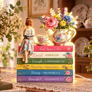 Può includere: Una lampada decorativa composta da una pila di sei libri colorati con accenti floreali e testi religiosi. Una statuina che legge è seduta sopra, accanto a una teiera e una tazza da tè a fiori. I libri riportano i titoli God Says I Am, Loved, Enough, Amazing, Strong e Beautiful.