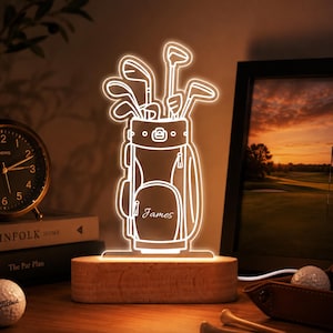 Può includere: Una luce notturna a LED personalizzata con un'illustrazione a linee bianche di una sacca da golf piena di mazze. La sacca presenta il nome James inciso sulla tasca anteriore. Il design in acrilico è montato su una base in legno rettangolare.