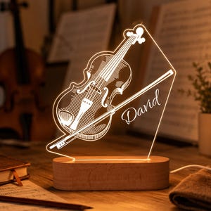 Op de afbeelding: Een gepersonaliseerd LED-nachtlampje met een acrylplaat waarop een viool en strijkstok zijn gegraveerd. De naam David staat in schrijfletters op het paneel. De lamp is gemonteerd op een rechthoekige houten voet met een stroomkabel aan de zijkant.