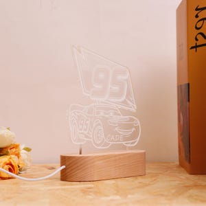 May include: A Lightning McQueen night light with a wooden base. The light features a white outline of the race car with the number 95 and the name CADE. The light is connected to a white cord.