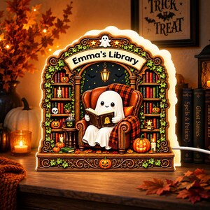 May include: An illuminated wooden display featuring a ghost sitting in a brown armchair reading a book. The scene includes bookshelves filled with colorful books, pumpkins, and a banner that reads Emma's Library. The design is arched with green vines.