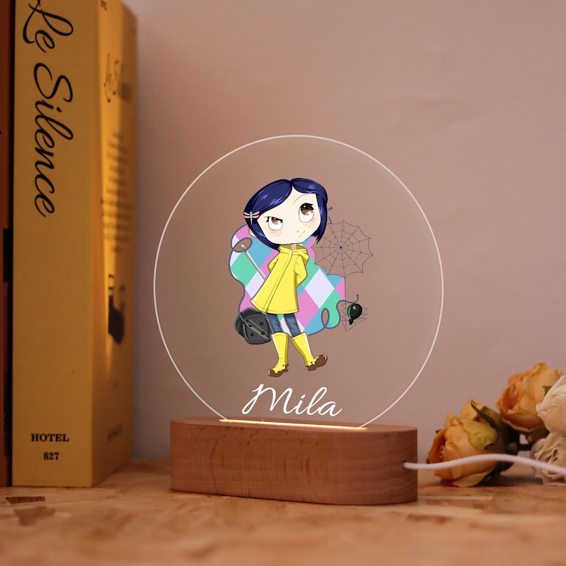 May include: A decorative night light featuring a cartoon character in a yellow raincoat, with the name "Mila" in script. The light has a wooden base and a clear, circular acrylic panel with the image. A book is visible in the background.